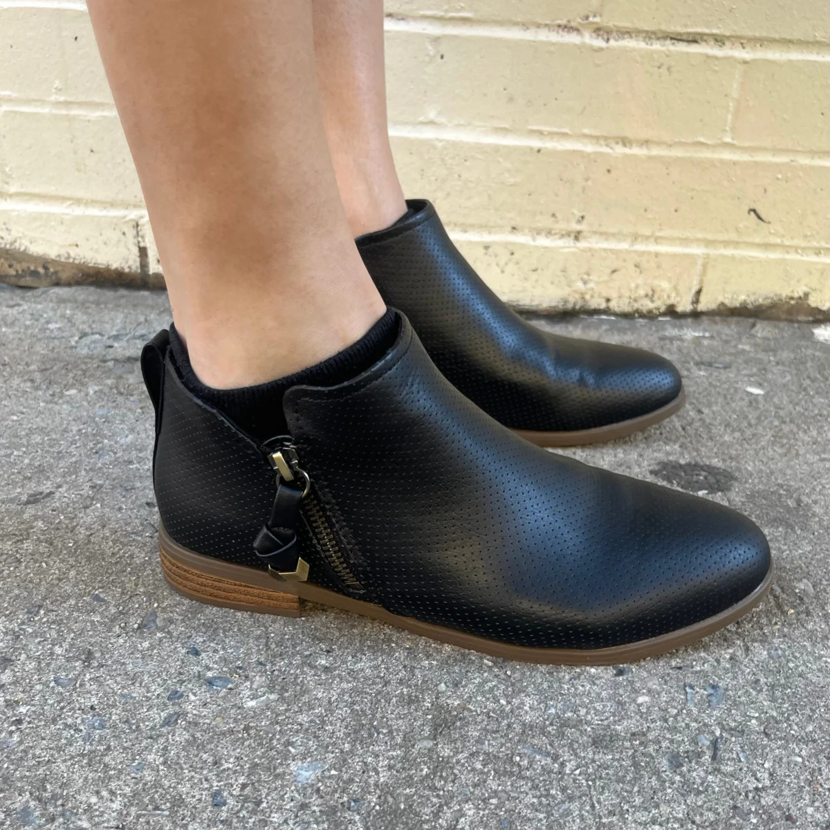 Black ankle boots with a perforated texture and side zipper, worn on a person standing on a textured surface. Black ankle boots with a perforated texture and side zipper, worn on a person standing on a textured surface.