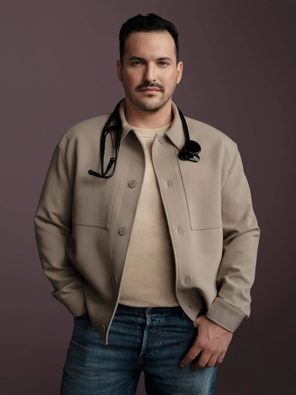 individual wearing a beige jacket with a stethoscope standing against a neutral background